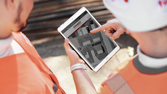 A photo showing the backs of two STRABAG employees on a construction site, looking together at a BIM model on a tablet.