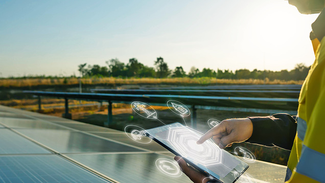 Photo of an employee in safety gear standing next to solar panels and operating a tablet.